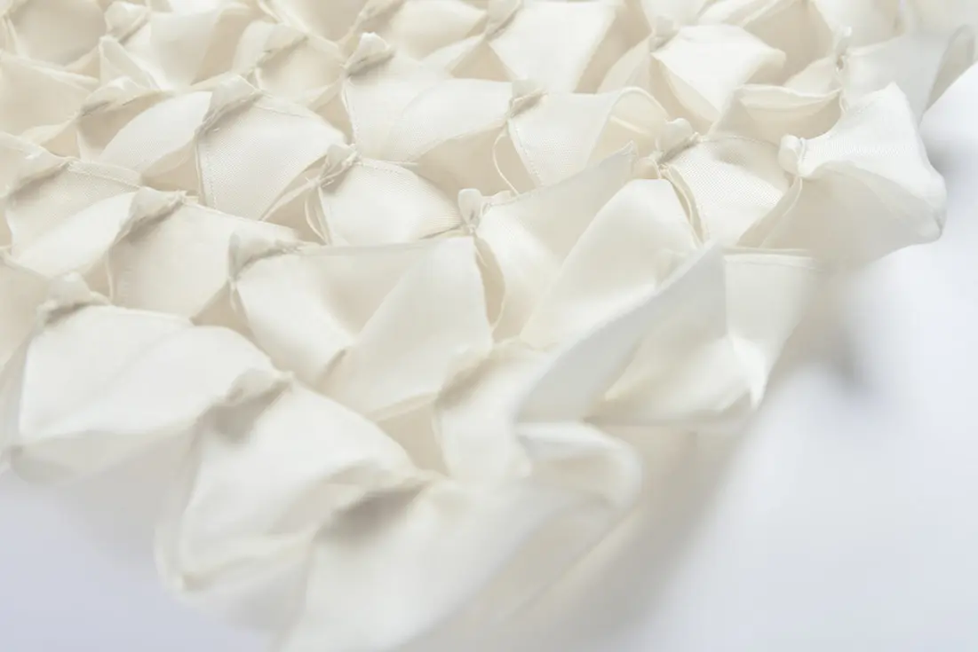 Cream-colored textile with a textured, three-dimensional surface formed by fabric manipulation techniques.
