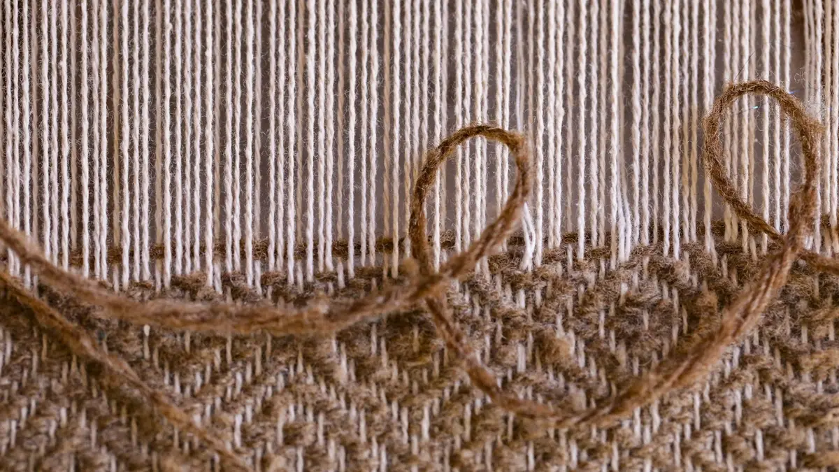 Close-up of beige handwoven textile showing intricate weaving patterns and threads on a loom.
