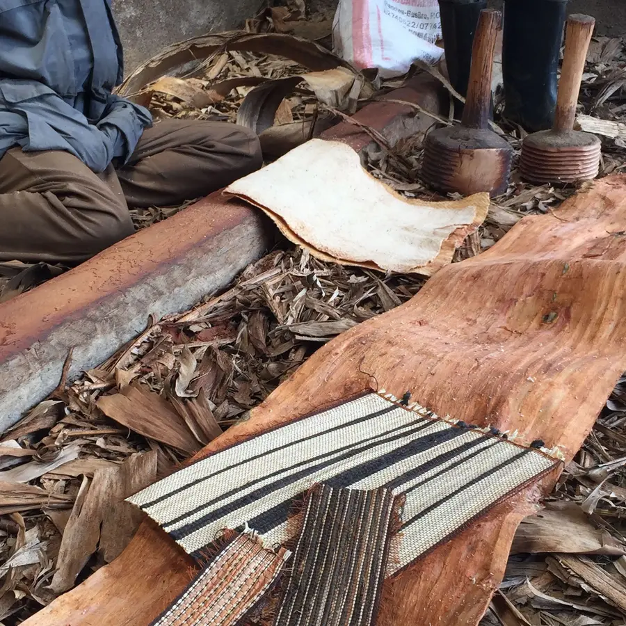 Bark cloth pieces and tools laid on a rustic surface, with a person seated nearby.