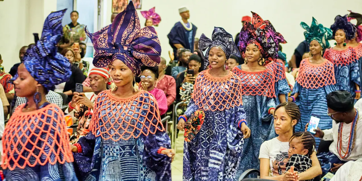 Women in colorful traditional garments with elaborate headwraps walk in a festive procession.